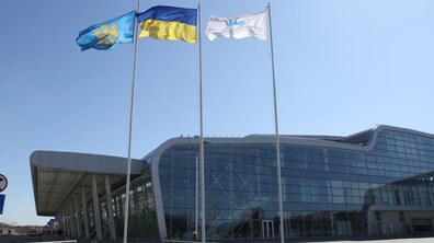 Термінал міжнародного аеропорту Львів. Велика скляна будівля, перед якою стоять три прапори: Львова, України та аеропорту «Львів»