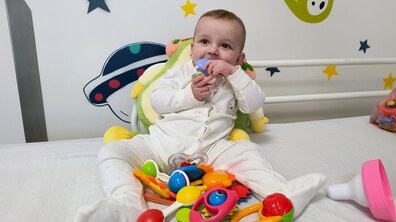 8-місячний Іванко сидить у дитячому ліжечку. Хлопчик одягнений у світлий комбінезон і тримає в роті невелику іграшку-прорізувач. Перед ним розкладено багато яскравих брязкалець та інших дитячих іграшок різних кольорів – червоного, жовтого, синього, зеленого. Позаду видно м’яку іграшку у вигляді тваринки, що підтримує дитину зі спини. Стіна оформлена веселими наліпками: зірками, ракетою та кометою.