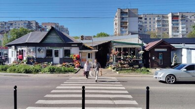 Міська вулиця, де двоє пішоходів переходять дорогу по зебрі. Позаду них невеликі одноповерхові магазини, зокрема квіткові крамниці та комерційні павільйони