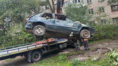 Занедбаний синій автомобіль, без вікон та розписаний графіті, завантажують на евакуатор. На тлі - цегляний будинок та зелені дерева.