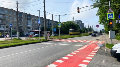 Міська вулиця з регульованим пішохідним переходом, який має яскраву червоно-білу розмітку. На другому плані видно багатоквартирні будинки, світлофори, дорожні знаки, а також приватний та міський транспорт, зокрема, автобус біля зупинки та жовтий трамвай, що рухається виділеною смугою біля зелених дерев.