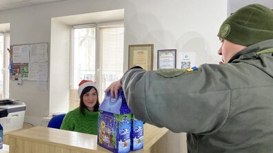 Військовий ставить на стіл два пакунки з солодощами. За столом сидить жінка.