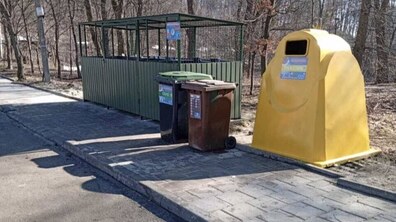 облаштований майданчик для роздільного збору сміття біля дороги. На вимощеній плиткою ділянці стоять кілька контейнерів: великий жовтий контейнер для пластику та два менші баки — зелений і коричневий. Позаду них розташований огороджений майданчик із металевим каркасом, а навколо видно дерева парку без листя.