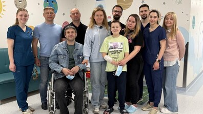 Спільне фото родини донора, реципієнта і медичних працівників дитячої лікарні святого Миколая та Центру трансплантології Львова. Усі дивляться в камеру та усміхаються. Позаду видно розмальовані стіни лікарняного холу.