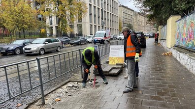 Вул. Личаківська, де розпочали ремонтні роботи на тротуарі. Два робітники у спецодязі демонтують старе покриття біля металевої огорожі. На задньому плані видно припарковані автомобілі, сучасні будівлі, дерева.