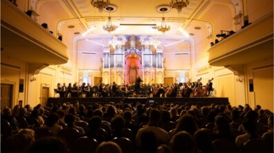 Виступ симфонічного оркестру у величній концертній залі з класичним інтер'єром у золотавих тонах. На сцені під керівництвом диригента грають музиканти, а на задньому плані височіє великий старовинний орган, підсвічений рожевим світлом. На передньому плані видно заповнений глядачами партер, де люди зі спини слухають музику та знімають подію на телефони.