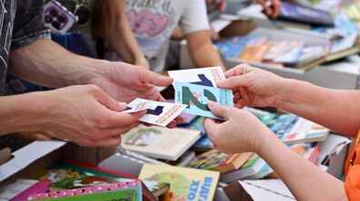 Учасники обміну тримають у руках картки з номерами та написом «Книжкова площа 2026». На задньому плані видно численні коробки й розкладені прилавки, заповнені різноманітними яскравими книжками.