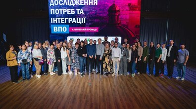 Групове фото учасників заходу на сцені. На задньому плані — великий екран із написом «Дослідження потреб та інтеграції ВПО у Львівській громаді».