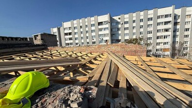 Облаштування тимчасового накриття у будинку на вул. Патона, 25 - розкладені дерев'яні балки та яскраво-жовта будівельна каска на передньому плані. На задньому плані розташований багатоповерховий житловий будинок зі світлими стінами та багатьма вікнами.