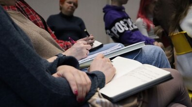 Група людей сидить в приміщенні. У їхніх руках записники та ручки