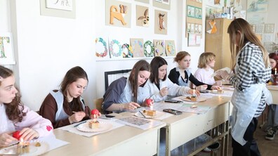 У світлій навчальній аудиторії група дівчат-підлітків сидить за довгим столом, зосереджено розписуючи великодні яйця. Перед кожною ученицею стоїть запалена свічка на дерев’яній підставці, за допомогою якої вони нагрівають писачки для роботи з воском.