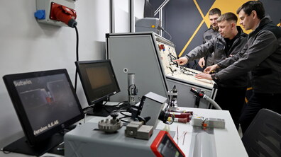 Троє працівників у формі стоять біля навчального стенда з електропроводкою та обладнанням і разом уважно розглядають, один із них показує рукою на деталі.
