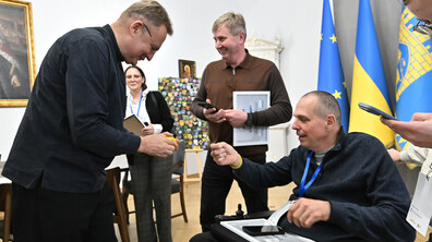 Нагородження сертифікатами учасників першої резиденції доступності. У світлій урочистій залі мер Львова Андрій Садовий нахилився до чоловіка у кріслі колісному, щоб почепити йому на руку браслет. Обоє усміхаються. На тлі — прапори України та ЄС, а поруч стоять люди з телефонами й відзнаками.