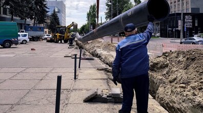 Вулиця Зелена під час заміни водопроводу. Робітник Львівводоканалу керує великою чорною трубою, яку екскаватор опускає в довгу траншею вздовж тротуару. Навколо — техніка, земляні насипи, будівлі та дерева під хмарним небом.
