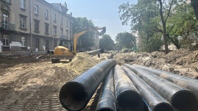 Ремонтні роботи на вул. Залізничній. На передньому плані лежать великі труби, поруч працює екскаватор і розкопана траншея для заміни підземних комунікацій. Обабіч вулиці видно житлові будинки та дерева.