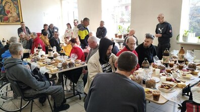 Світла кімната з великими вікнами, де за довгими столами сидять люди — переважно чоловіки, є й кілька жінок. Вони їдять і спілкуються під час Великоднього сніданку. На столах — паски, яйця, хліб, гарячі страви та напої. Ліворуч видно чоловіка на інвалідному візку, у центрі стоїть чоловік у яскравій куртці. На стіні — ікона Богородиці.