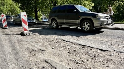 Вулиця Винниченка з пошкодженим асфальтом і ямами на передньому плані. Дорогу частково огороджено червоно-білими стовпчиками. Повільно їде позашляховик, позаду — інші авто. Праворуч тротуаром іде жінка.