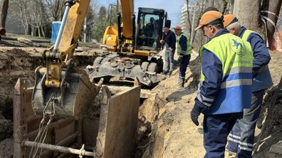 Ремонтні роботи на водопровідній мережі. У викопаній траншеї працює жовтий екскаватор, поряд встановлені металеві щити, які укріплюють стінки ями. Біля краю траншеї стоять кілька працівників у робочому одязі та світловідбивних жилетах із написом «Львівводоканал» і спостерігають за ходом робіт. Довкола видно дерева та міські будівлі, що вказує на проведення робіт просто вздовж міської вулиці.