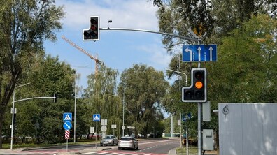 На міській вулиці видно світлофори, дорожні знаки та розмітку. Дорогою рухаються два автомобілі, навколо — зелені дерева