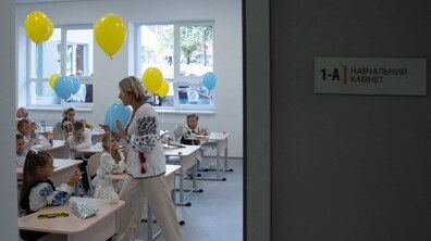Вчителька розмовляє з дітьми у класі, які сидять за партами. біля них блакитні і жовті надувні кульки. поряд на стіні напис – 1-А навчальний кабінет. Фото ілюстративне.