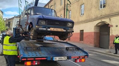 На брукованій вулиці евакуатор піднімає на платформу синій автомобіль марки «Москвич» за допомогою ременів та крана. Робітник у жовтому світловідбиваючому жилеті допомагає завантажувати машину. Поруч – міська будівля