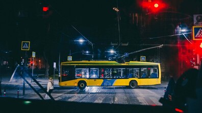 Трамвай їде нічним містом у світлі ліхтарів та червоному світлі світлофора. Перед ним жінка йде пішохідним переходом.