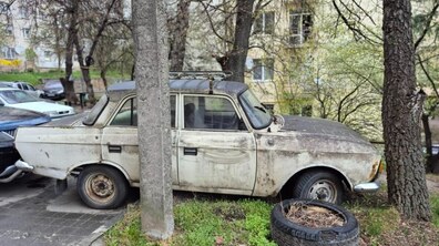 Покинутий старий світлий автомобіль, покритий брудом. На другому плані частково видно дерева, будинки та інші автомобілі.