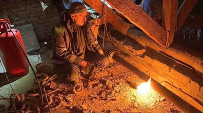 Чоловік, на горищі будинку, одягнений у темний зимовий одяг, нахилився до металевої труби і тримає спеціальний пристрій для нагрівання. З пальника виходить яскраве полум’я, яким він розморожує замерзлу трубу. Навколо видно дерев’яні балки даху та стару цегляну стіну. Простір горища вузький і напівтемний.