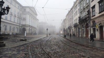 Площа Ринок у Львові, оповита густим туманом: брукована дорога з трамвайними коліями тягнеться вперед і зникає в тумані; з обох боків стоять старовинні багатоповерхові будівлі зі світлими фасадами, арками та балконами, зокрема зліва є будівля міської Ратуші. Вздовж вулиці є ліхтарі, зліва стоїть інформаційний стенд, а праворуч на будинках видно кілька синьо-жовтих прапорів. Вулицею йдуть поодинокі пішоходи в темному одязі.
