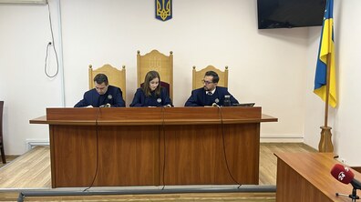 У залі суду троє суддів у темно-синій формі сидять за дерев’яним столом на тлі герба України. Перед ними розміщені мікрофони та робочі матеріали.