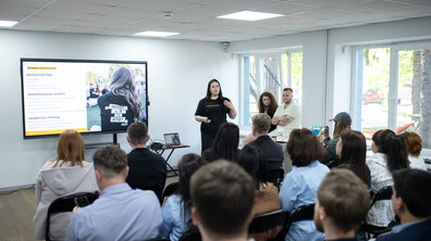 Презентація проходить у світлій сучасній аудиторії. Спікерка — дівчина у чорній футболці — стоїть біля великого екрана з текстом та інфографікою українською мовою. Слухачі сидять на стільцях спиною до камери, уважно спостерігаючи за виступом.