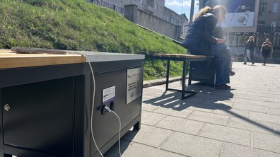 На лавці з сонячними батареями заряджається телефон. Поруч на сусідній лавці сидить жінка. Позаду видно перехожих