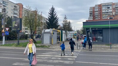 На міській вулиці під похмурим небом люди переходять пішохідний перехід біля ряду невеликих кіосків і житлових будинків