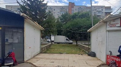 Дві невеликі тимчасові споруди, між якими прохід викладений плиткою. Позаду розташовані дерева, дорога та багатоповерховий житловий будинок