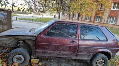 Покинутий автомобіль вишневого кольору, у якого відсутня частина переднього крила, тому видно пружину підвіски біля переднього колеса. Машина стоїть під деревом серед опалого осіннього листя біля житлового будинку.