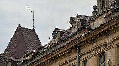 Верхня частина будівлі з пошкодженим дахом внаслідок російської атаки. Частина покрівлі зруйнована, видно уламки та обгорілі елементи, з-під яких пробивається невелике полум’я, а також деякі нависаючі аварійні елементи. Небо сіре й похмуре, що підсилює відчуття руйнування і тривоги.