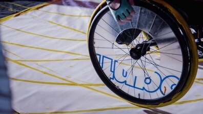 Крупний план колеса крісла колісного, що рухається по розмальованому білому полотні, на якому видно жовті лінії та частину синього напису.