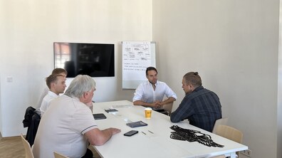 За великим білим столом сидять чоловіки і розмовляють. Перед ними телефони та ручки. Позаду на стіні висить телевізор і розташована дошка, на якій є намальовані схеми чорним маркером