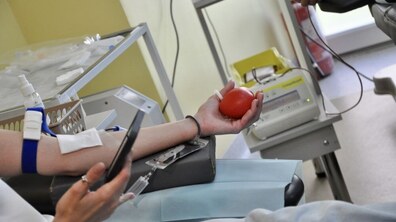 Людина здає кров, сидячи у кріслі. У одній руці тримає червоний мячик, а у іншій - телефон. Довкола медичне обладнання для здачі крові