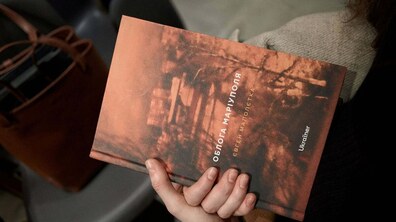 Людина тримає в руках книгу з назвою «Облога Маріуполя» Євгена Малолетки. Обкладинка виконана у темних, теплих тонах.