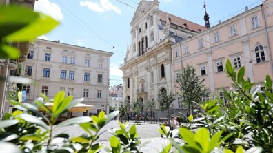 Знімок зроблено крізь зелене листя кущів. У центрі височіє Гарнізонний храм св. апп Петра і Павла. Його фасад оздоблений декоративними деталями та високими арковими вікнами. Над дахом піднімається темний шпиль. Площа вимощена світлою бруківкою. Видно молоді дерева, лавки та людей.