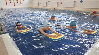 Група дітей займається плаванням у басейні з блакитною водою. Хлопці та дівчата в різнокольорових плавальних шапочках (блакитних, синіх, червоних) тримаються руками за жовто-сині дошки для плавання, відпрацьовуючи рухи ногами. Стіни басейну прикрашені яскравою мозаїкою у формі різнокольорових ромбів