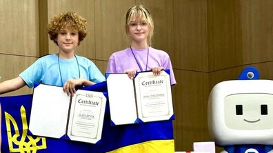 Анна Телегіна та Олесь Летита на сцені після вручення нагород. Школярі тримають у руках сертифікати та прапор України. Позаду на підлозі стоять подарункові коробки та велика фігурка робота.
