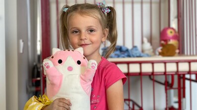 Дівчинка в рожевій футболці з хвостиками та блакитними бантиками тримає правою рукою, на якій апарат зовнішньої фіксації та жовта пов’язка, м'яку іграшку у вигляді кота. Позаду неї видно дитяче ліжечко з іграшками