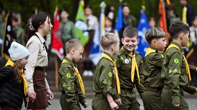 Хлопці-пластуни, одягнені в зелені сорочки з нашивками та жовті хустки, йдуть разом у строю, а поруч із ними іде їхня наставниця. На задньому плані видно інших пластунів та різнокольорові прапори різних куренів та підрозділів Пласту. Кадр із урочистостей з нагоди 114-ї річниці з Дня першої Пластової присяги у Львові.