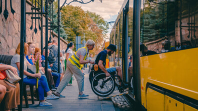 Зупинка громадського транспорту. У центрі кадру чоловік на кріслі колісному, за допомогою водійки, заїжджає до низькопідлогового жовтого трамвая спеціальним відкидним пандусом.