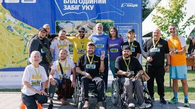 Учасники Першого благодійного забігу УКУ, які зібралися біля інформаційного стенда заходу. Серед учасників — ветерани, люди на кріслах колісних, військові та цивільні, які разом долучилися до благодійної ініціативи. Багато з них мають медалі та стартові номери, а дехто — протези чи засоби реабілітації.