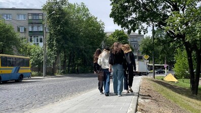 Четверо дівчат йдуть по новому вимощеному плиткою тротуару вздовж брукованої вулиці. Навколо ростуть зелені дерева, видно житлові будинки, автобуси та автомобілі