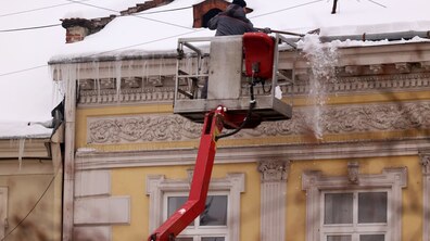 Чоловік, піднятий автовишкою до рівня даху будинку, чистить покрівлю від снігу і буруль