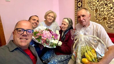 Ювілярка, її родичі та працівник управління соціального захисту усміхаються, позуючи для спільного селфі. Пані Віра тримає в руках великий букет різнокольорових квітів, а чоловік поруч – великий кошик з фруктами.